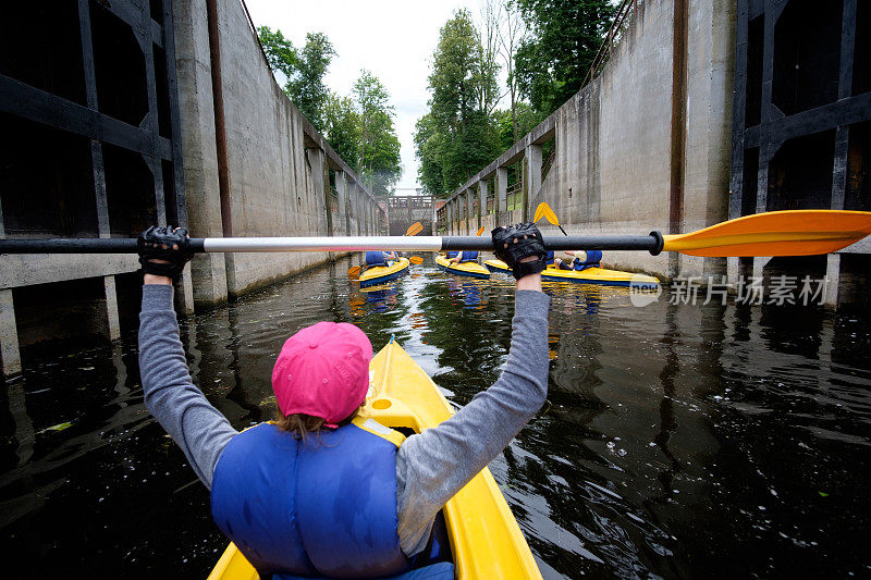 小女孩戴着粉红色的皮划艇帽在河道上划水闸，从后面看，皮划艇队