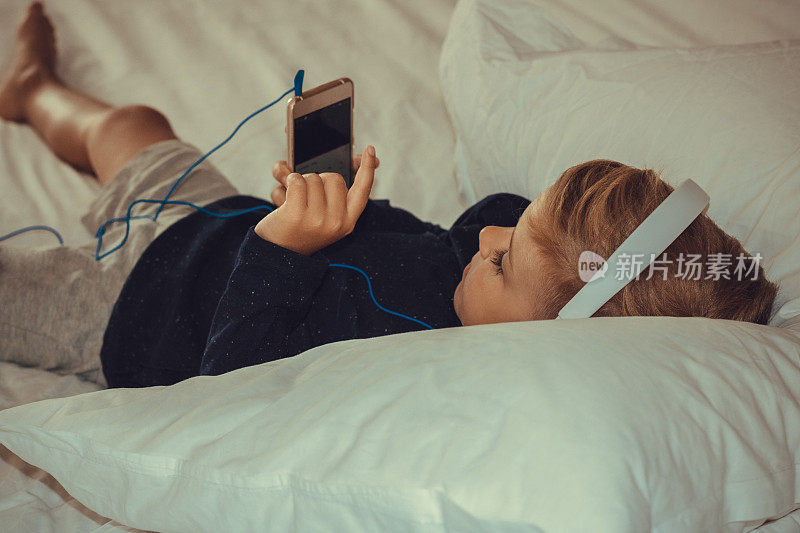 放松的男孩使用智能手机和听音乐在床上。