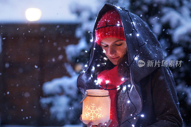 美丽的黑发少女穿着连帽大衣，同时在一个下雪的冬天晚上在户外举着圣诞灯