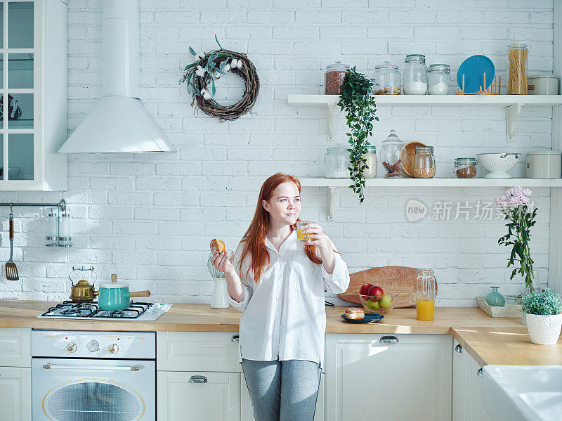 红色头发的年轻女子，在厨房里吃着自制的糕点，喝着橙汁当早餐，做着白日梦