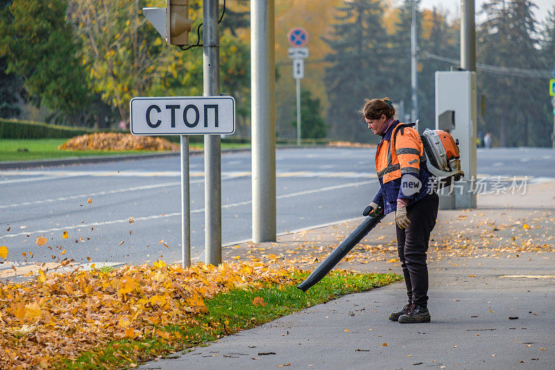 一名女公用事业工人在用吹风机清除公园巷子里的落叶。