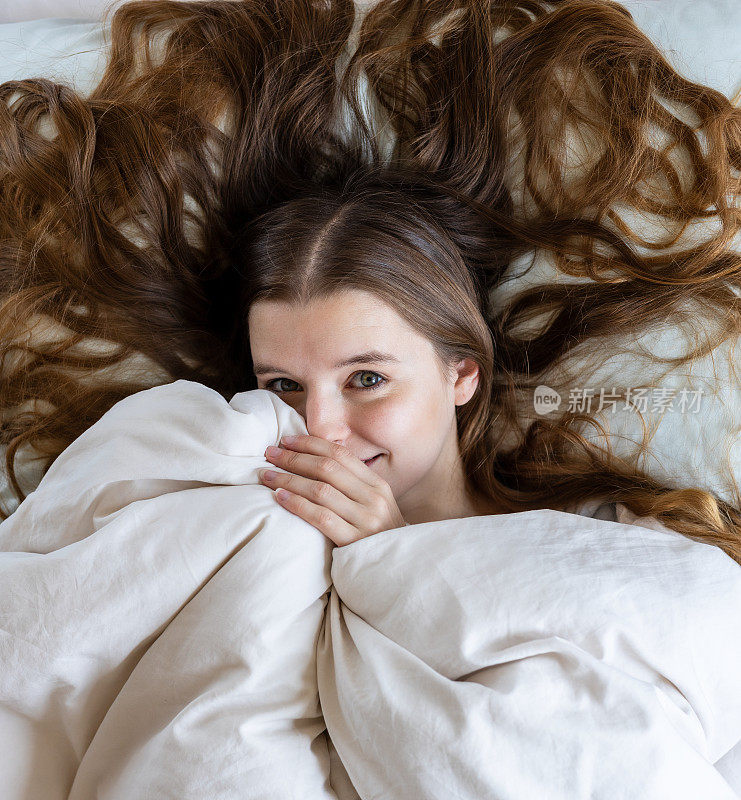 一个害羞迷人的金发年轻女子在睡床上披着羽绒被，嬉闹着诱惑着她，躲在被子下面