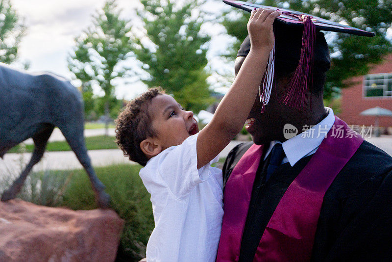 非裔美国男大学毕业生戴着帽子和长袍，在毕业典礼上和他三岁的儿子玩
