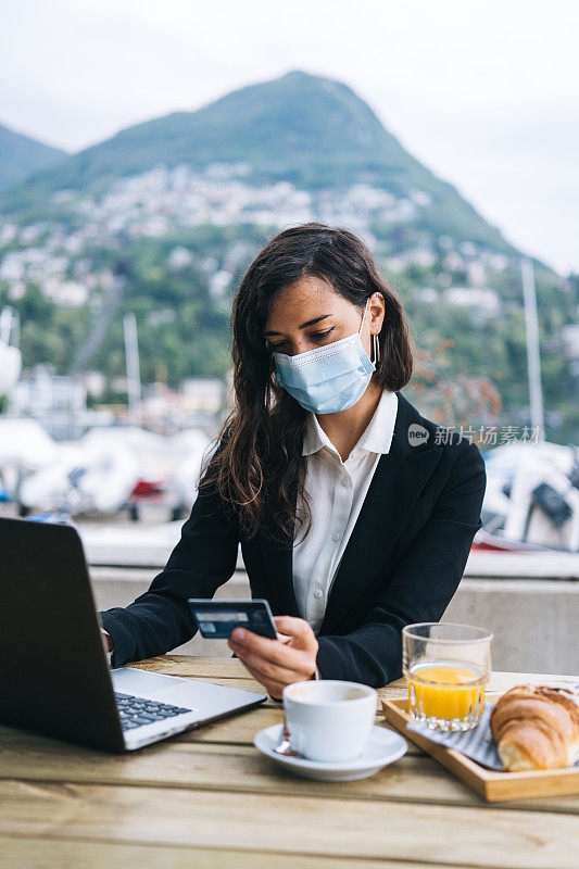 年轻的商业女性在户外咖啡馆使用信用卡，戴着防护面罩