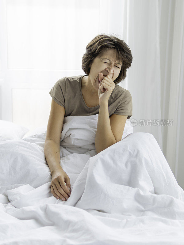 困倦的女人盖着白毯子在床上打哈欠。早上很难早起。