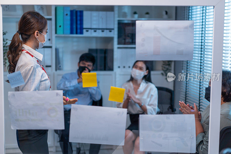 一群亚洲商人和妇女戴着口罩，在办公室开会。员工群策群力，以团队的形式工作，在纸上逐点计划和讨论项目。现代同事或同事的公司。