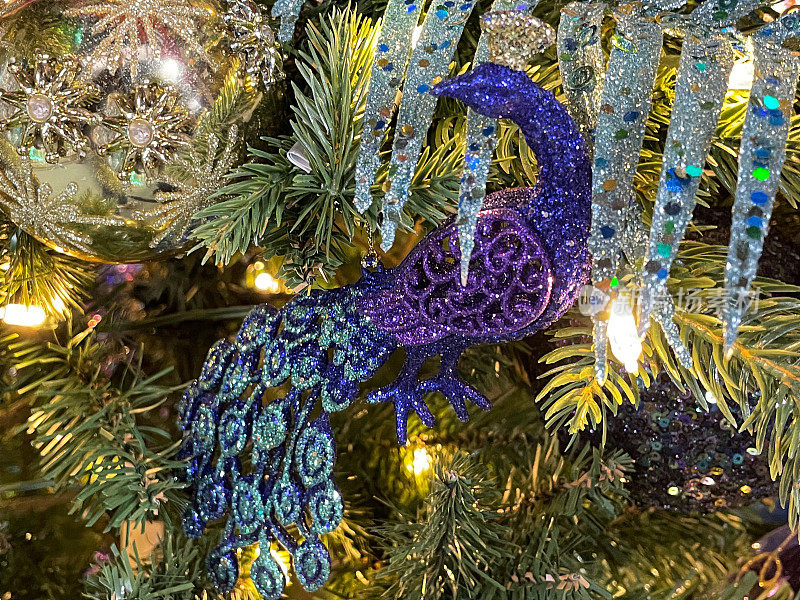 特写图像的个人，蓝色和紫色闪光孔雀装饰人造圣诞树树枝，黄金珠宝小饰物，云杉针，模糊的白色精灵灯背景