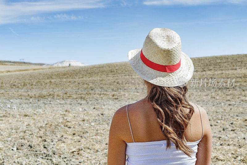 在炎热的夏日午后，戴着草帽的年轻女子从后面望着田野和远处的山