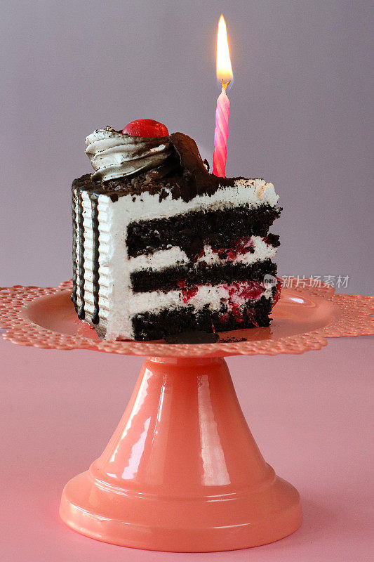 黑森林蛋糕片的图像，三层蛋糕层被橙色蛋糕架上的鲜奶油隔开，点燃的生日蜡烛，管状鲜奶油花环，莫雷罗樱桃，豪华巧克力蛋糕，巧克力刨花，粉紫色的背景，重点在前景