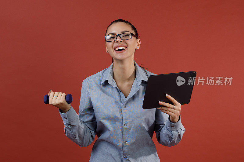 年轻的女商人在蓝色衬衫暖哑铃在办公室，而工作在平板电脑。多任务业务和时间管理。