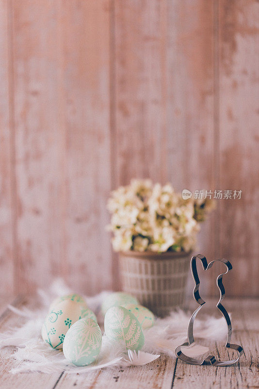 可爱的近距离自我绘制的薄荷绿色复活节彩蛋和白色羽毛，复活节兔子饼干切割机和木色背景上有白色花朵的罐子