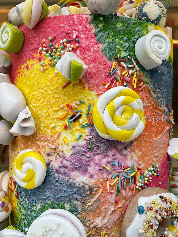 彩虹彩色冰冻生日蛋糕表面的特写图像，装饰有彩色糖屑和翻糖糖霜形状的糖果和糖果，集中在前景