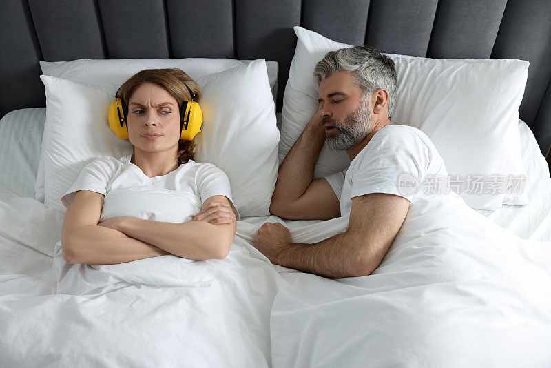 恼怒的女人戴着耳机躺在家里打鼾的丈夫旁边