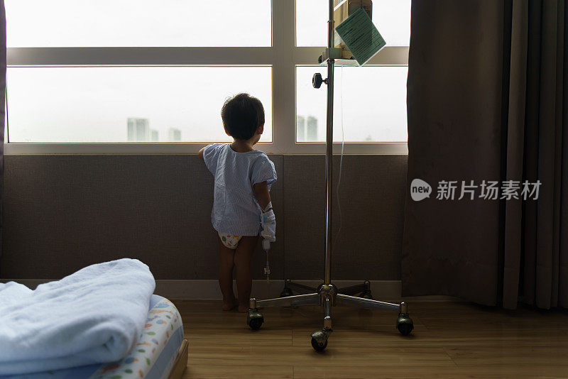生病的小男孩站在医院的后视图，透过窗户向外看。