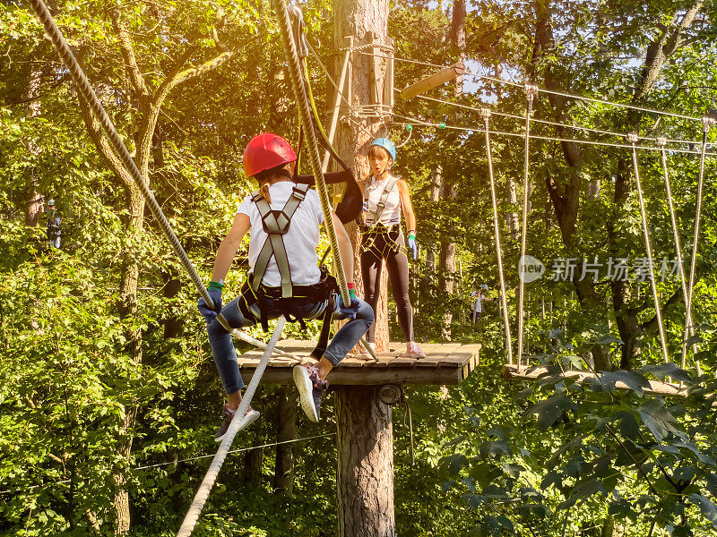 夏日里，一个十几岁的女孩穿着安全装备和头盔，在一个攀岩冒险公园里沿着树间的索桥散步。木林中的绳索公园。冒险公园特写。夏季的乐趣和运动冒险的人。