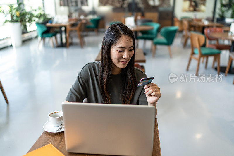 一个亚洲女人坐在咖啡店里，拿着信用卡，用着笔记本电脑