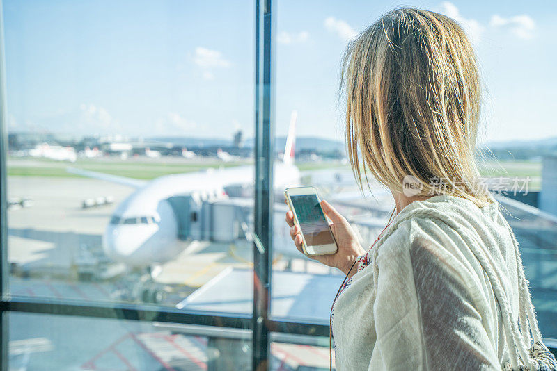 在机场等待航班的年轻女子坐在长凳上，手里拿着手机和护照