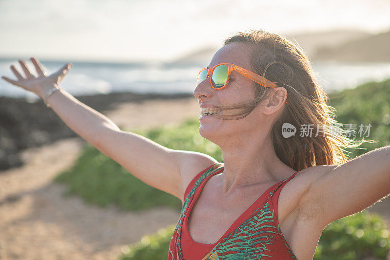 夕阳西下，拥抱大自然的快乐年轻女子;女人站在沙滩上，伸开双臂。