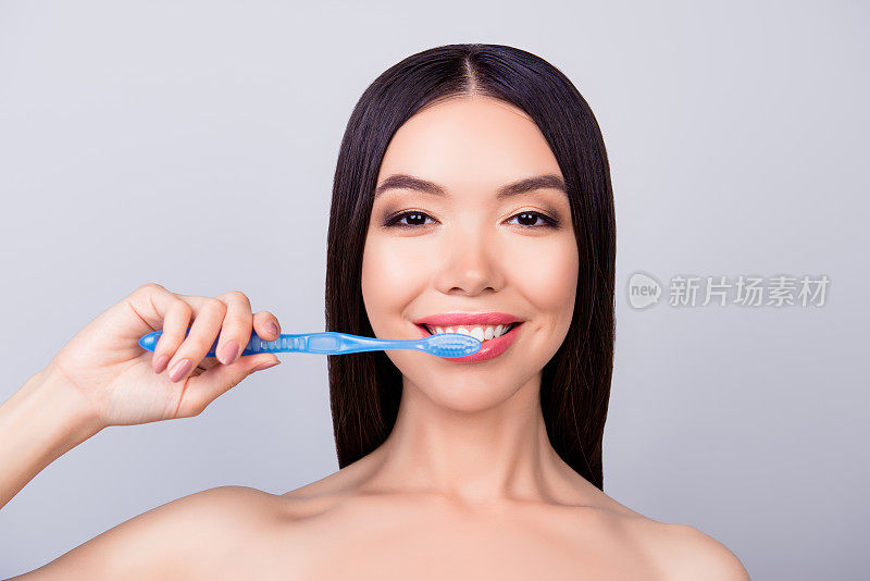 牙齿健康的概念。美丽的亚洲女孩正在清洁她的牙齿与蓝色的牙刷，在浅灰色的背景，她有灿烂的微笑