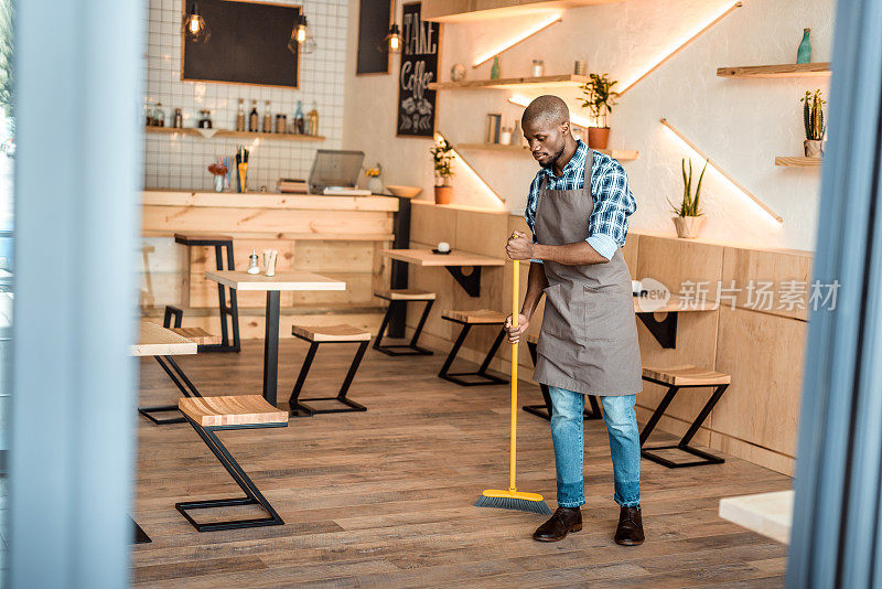 英俊的非裔美国工人在咖啡店用扫帚扫地