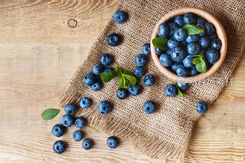 新鲜采摘的多汁和新鲜的蓝莓与绿色薄荷叶在轻木碗上帆布黄麻布古董乡村桌子。越桔水平木制背景。健康饮食和抗氧化营养概念。俯视图，复制空间。