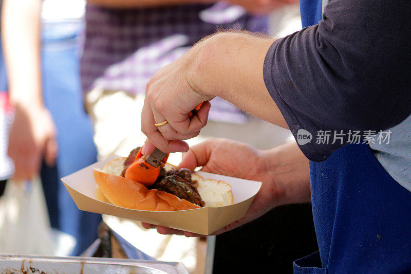 这是一幅自制外卖切碎的烤牛排三明治食谱，厨师用热狗香肠卷在烧烤街的小吃摊上吃着，手里拿着装着沙拉、洋葱和芥末酱的纸盒，这就是快餐