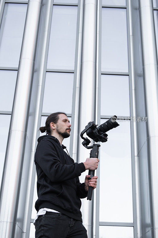 年轻的专业摄像师手持专业摄像机，安装在三轴框架稳定器上。专业设备有助于制作高质量的视频而不摇晃。一个穿着白色连帽衫的摄影师正在录像。