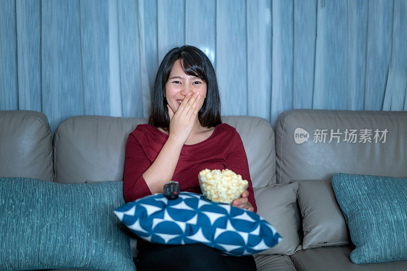 年轻的亚洲女性看电视悬疑电影或新闻，看起来开心和有趣，吃爆米花深夜在家客厅的沙发上隔离。