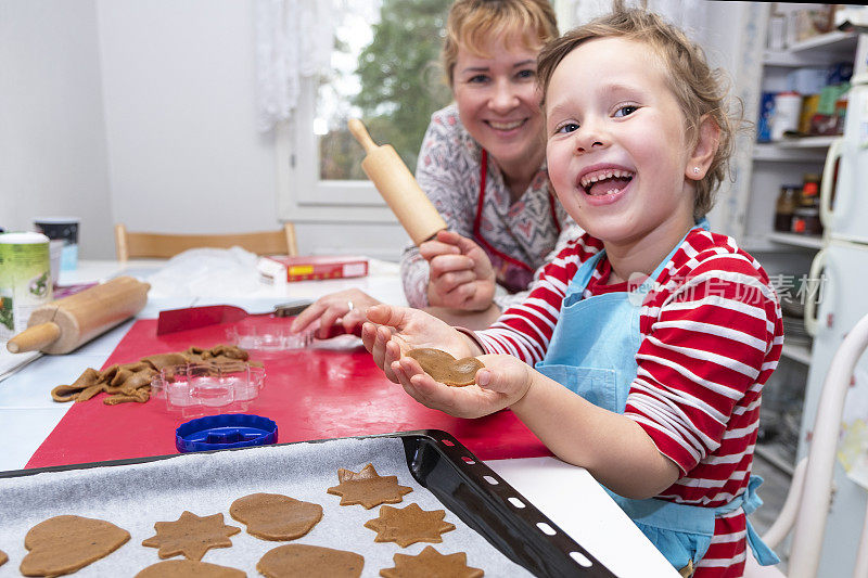 一个年轻的白人妇女和一个5岁的女孩在家庭厨房里烤姜饼。那孩子高兴地笑着。