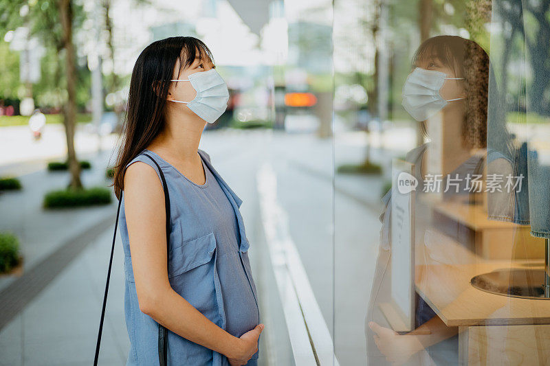 亚洲孕妇戴着口罩在城市里走着，看着商店橱窗