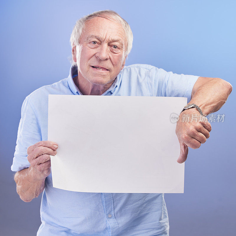 摄影棚里，一位年长的男子举着一块空白的牌子，蓝色背景下的大拇指朝下