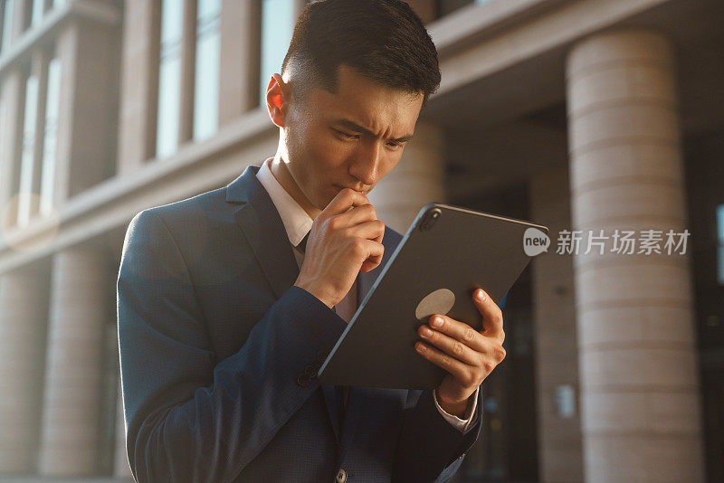 一位身穿蓝色西装、注意力集中的亚洲商人正在思考他在平板电脑上收到的一封电子邮件中描述的问题