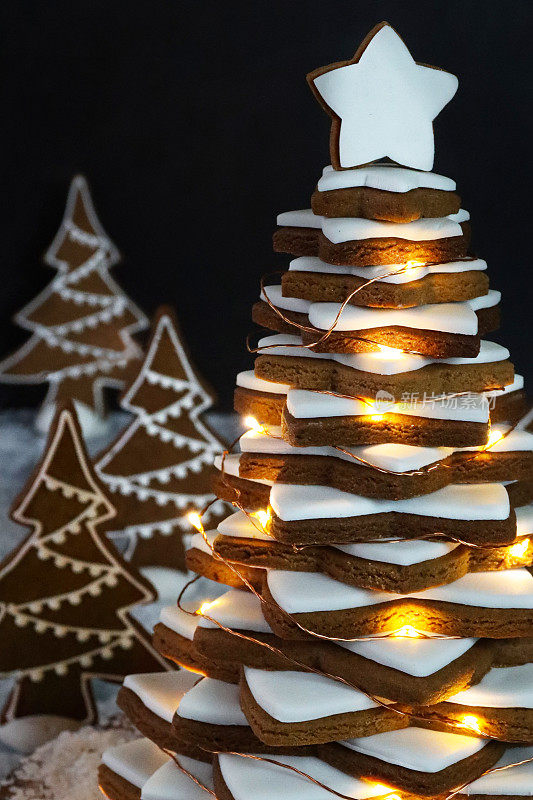近距离拍摄的圣诞树，在木制蛋糕架上堆放着覆有白色皇家糖霜的姜饼星饼干，装饰着发光的仙女灯，针叶林场景与圣诞树饼干，人造雪，黑色背景