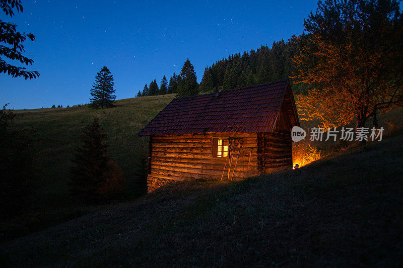 传统的乡村山间木屋在夜晚被温暖的火把和火照亮。山中的蓝色时光。夜景图像与草地和高大的冷杉树。