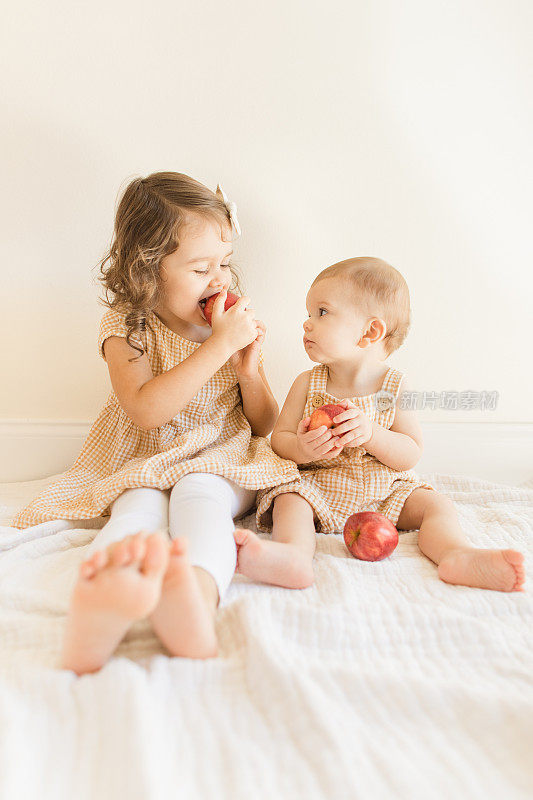 2022年秋天，一个3岁的蹒跚学步的女孩和她9个月大的小弟弟穿着配套的黄色格纹衣服，在家里地板上的奶油色薄纱毯子上玩和吃新鲜的红色节日苹果