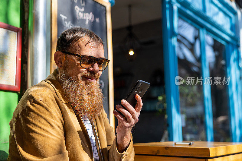 留着长胡子的城市白人男子，坐在路边咖啡店里用手机