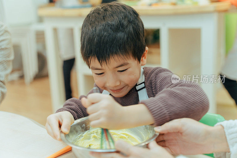 年轻的十几岁快乐的亚洲男孩混合饼干面团在碗里烤饼干，而坐在桌旁的室内