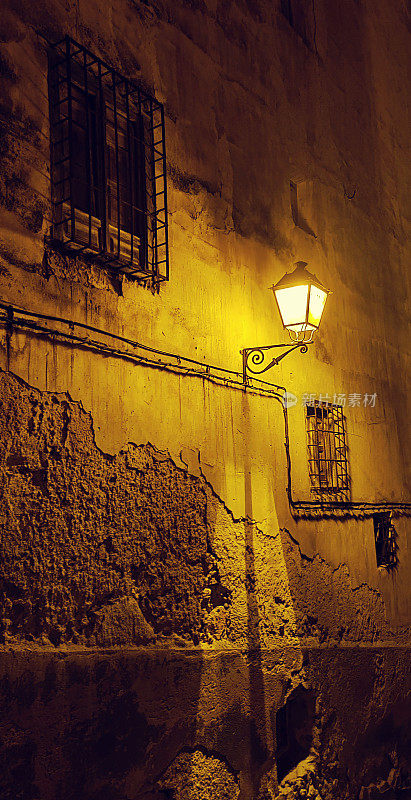 夜晚的老城区，一面破旧、肮脏、破碎的墙的正面，有金属窗户，还有一盏古老的路灯，发出微弱的橙黄色灯光。黑色垂直明信片令人毛骨悚然的壁纸背景