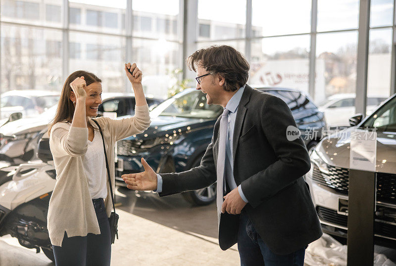 在一家汽车经销商的室内，一位女顾客和一名男性汽车销售员并排站在一起，在做了一笔大交易后，她举起双臂，露出露齿的微笑