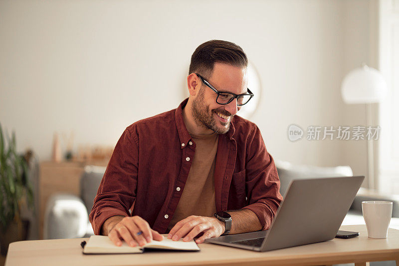 微笑的商人在家办公，做着笔记，看着笔记本电脑。