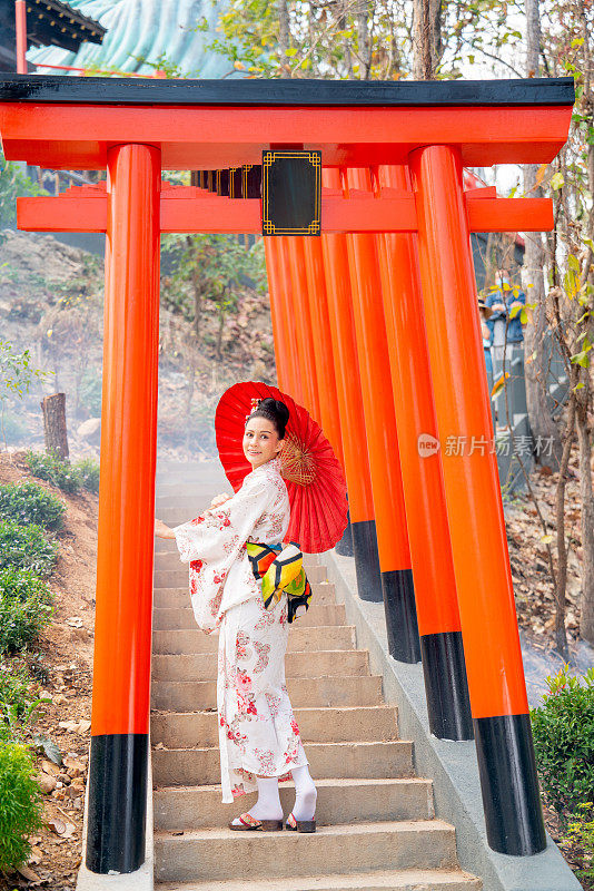 在泰国旅游景点的红色拱门入口处，穿着日式服装的漂亮女子手持红色雨伞，背对着镜头。