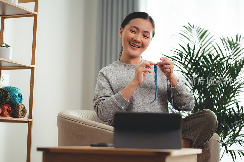 亚洲女性坐在家里的客厅里，看着如何在平板电脑上编织视频剪辑，享受着自己的爱好。