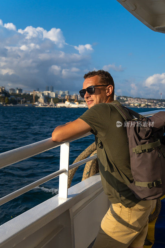 在一个阳光明媚的日子里，在伊斯坦布尔的渡轮上，戴着太阳镜、背着背包的快乐英俊的年轻男性游客享受着大海和这座城市。旅游的概念，旅游。垂直图