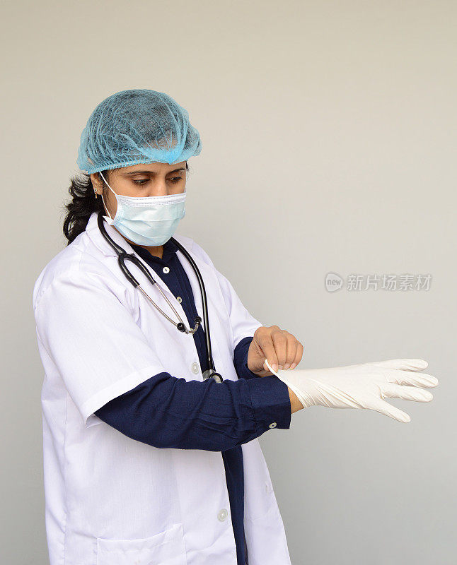 一名女医生或女医生的垂直照片，一脸茫然的表情，深蓝色的衬衫，白色的实验室外套，帽子，听诊器，准备戴上手术手套，在灰色的背景上，文字的复制空间
