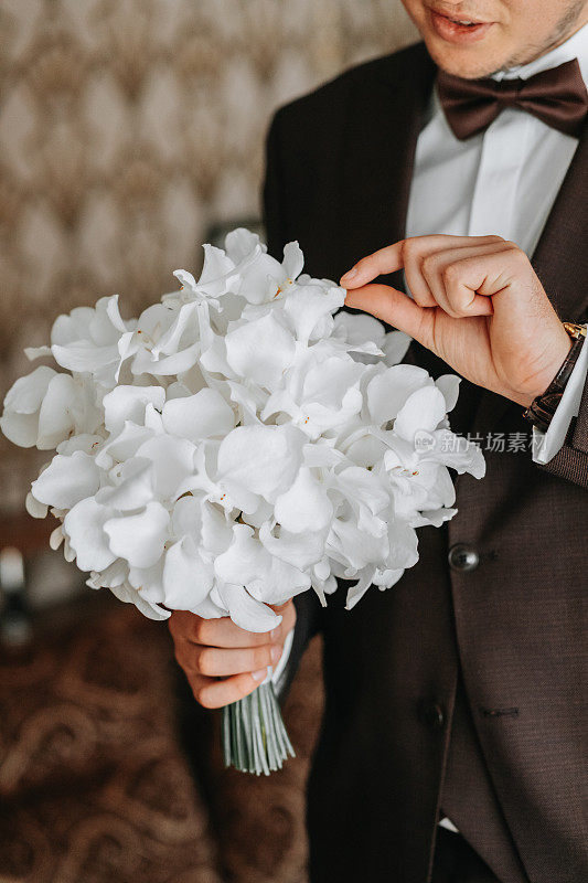 新郎穿着棕色夹克和白色衬衫，手捧一束花，房间里的装饰非常漂亮。新郎看着捧花
