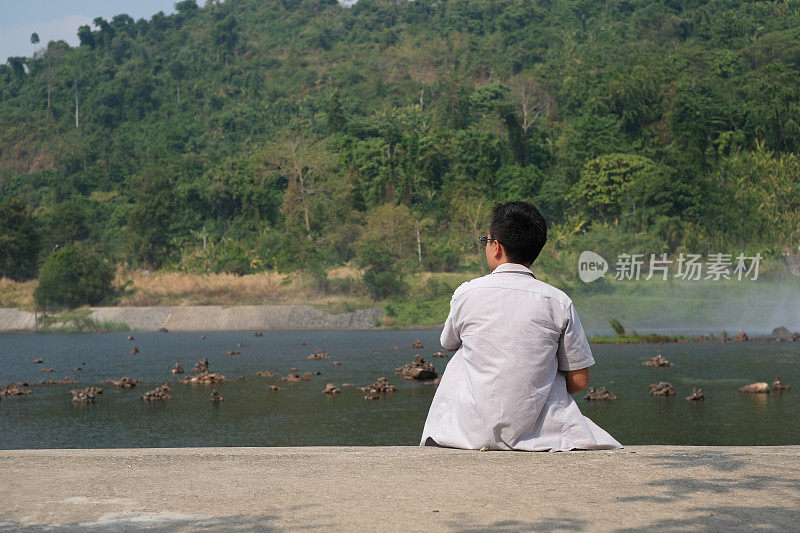 年轻的亚洲男子望向远方，坐在河上的地板上，表现出自由，以人类的责任观念对自然保持绿色。