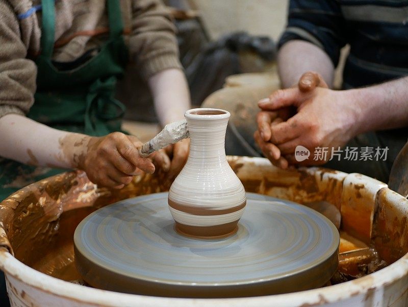 陶工师傅帮助孩子在一个电动驱动的现代陶工车轮上用白色油漆画一个陶罐。在陶瓷花瓶上画图案。一个孩子和一个成年人的手都沾满了泥土。