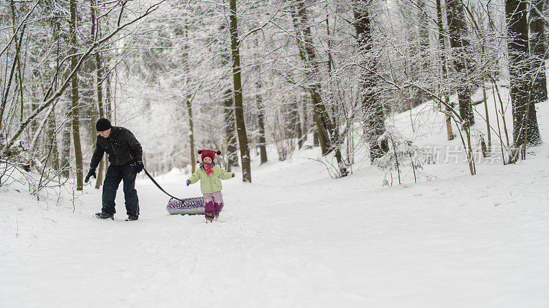 联系是老年人快乐和活动的重要关键。70岁的老人，爷爷，走在他2岁的孙女在冬天的森林里的路上，拿着雪管。