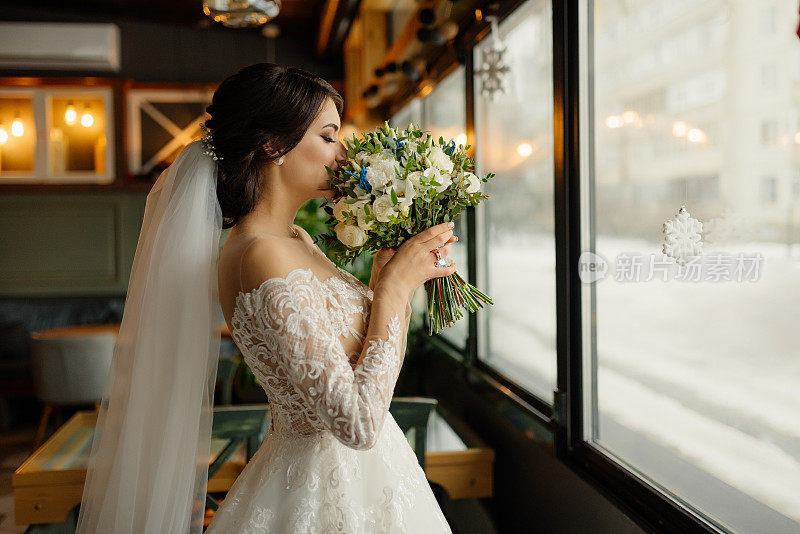 幸福的新娘闭着眼睛站在阁楼内部靠近窗户的地方，闻着婚礼花束。身穿白色连衣裙，手持一束鲜花的漂亮年轻女子。新娘婚礼肖像