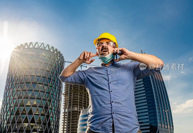 戴着黄色安全帽和N95防护面具的年轻聋哑工程师在现代商业建筑和建筑工地前使用智能手机